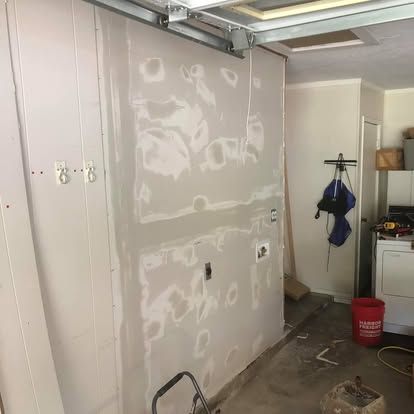 A garage wall with unfinished drywall, showing taped seams and joint compound patches, with a doorway visible in the back.
