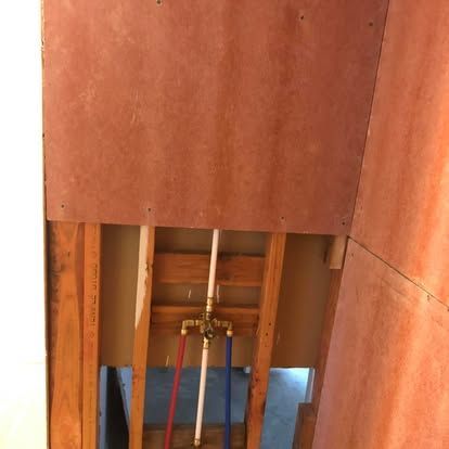 A brass shower mixing valve is mounted between wall studs with red, blue, and white PEX plumbing lines attached.