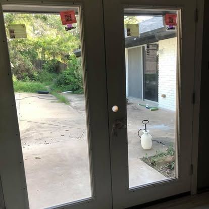 A view through glass double doors showing a concrete patio with a white spray canister and overgrown greenery outside.