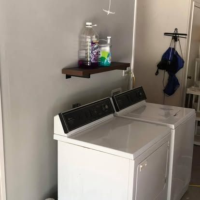 A laundry room with two white washing machines side-by-side below a dark wood shelf holding bottles of cleaning supplies.