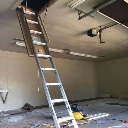An aluminum pull-down ladder is extended from a ceiling hatch into a dimly lit, unfinished garage space.
