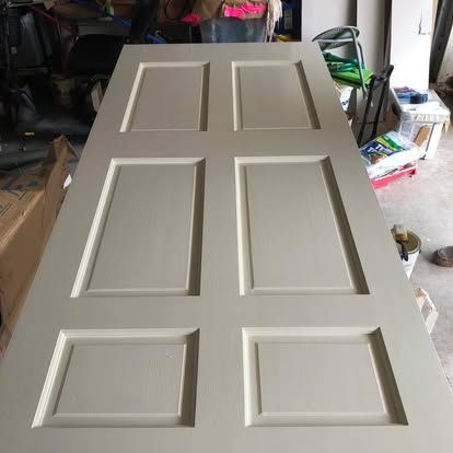 A light gray, six-panel interior door lies flat on a workshop floor, surrounded by painting supplies and equipment.