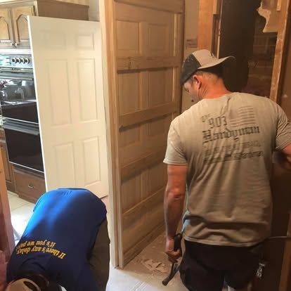 Two workers in a residential setting install a wooden door frame, with one working on the floor and one holding a hammer.