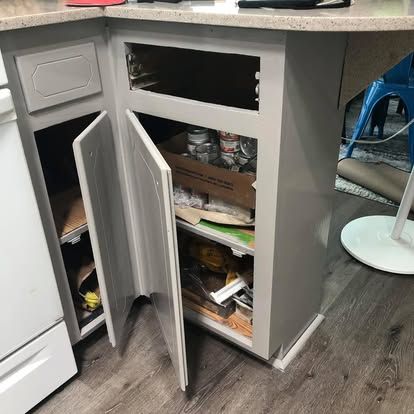 Gray corner kitchen cabinets with open doors, showing interior shelving and a missing top drawer face.