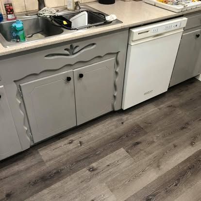 A kitchen sink area with light gray, decorative cabinets, a white dishwasher, and gray wood-look laminate flooring.