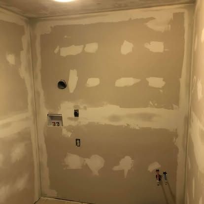 Unfinished drywall in a laundry area with patched screw holes and visible utility hookups on a beige wall.