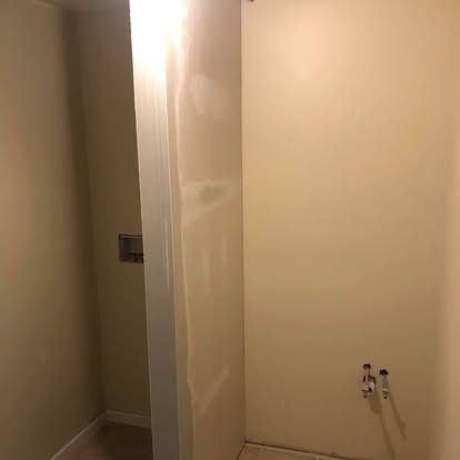 An unfinished interior corner wall with beige paint, exposed plumbing for a sink, and an open doorway to the left.