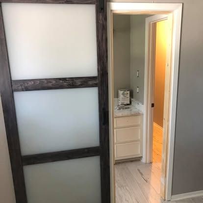A sliding dark wood barn door with three frosted glass panels stands partially open to a bathroom with a white vanity.