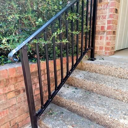 Black metal railing along a set of concrete steps next to a brick wall and leafy green bushes.