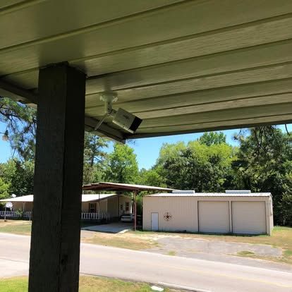 A white security camera mounted to a porch ceiling looking out toward a rural house and a metal workshop across the street.