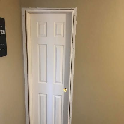 A white six-panel door installed in a tan wall, surrounded by unfinished white trim and visible nail holes.