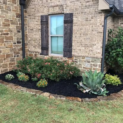 A stone house exterior features a window with dark shutters above a garden bed with shrubs, succulents, and black mulch.