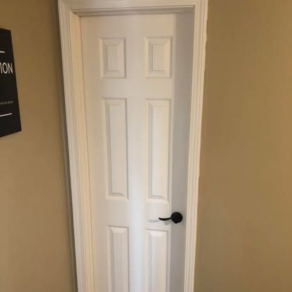 A white six-panel interior door with a black handle, set in a beige wall.