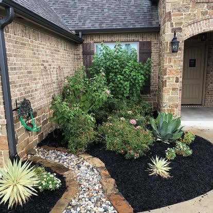 A landscaped house corner features a gravel path bordered by bricks, dark mulch, and various green shrubs and succulents.