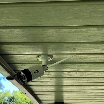 A white, bullet-style security camera with a Wi-Fi antenna mounted to a beige horizontal-paneled outdoor ceiling.