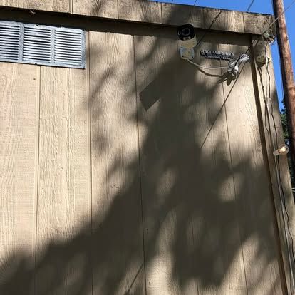 A security camera mounted on the side of a tan wooden building near a vent and a utility pole.