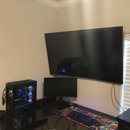 A computer setup with a gaming PC, a small monitor, and a large wall-mounted TV on a desk with a colorful mouse pad.