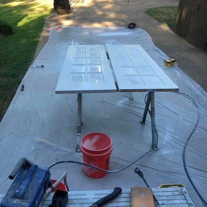 Two white paneled doors rest on sawhorses over a plastic drop cloth on a patio, with a paint sprayer and bucket nearby.