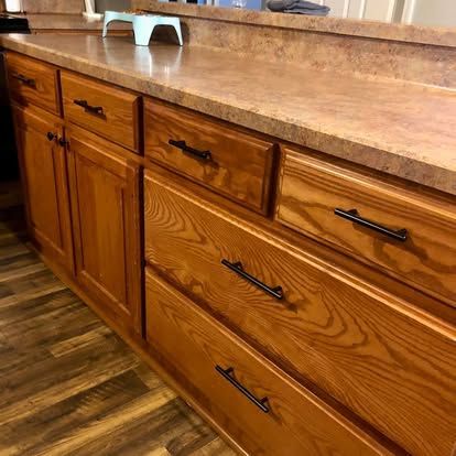 Medium-toned wooden kitchen cabinets with black handles, a speckled laminate countertop, and wood-grain vinyl flooring.