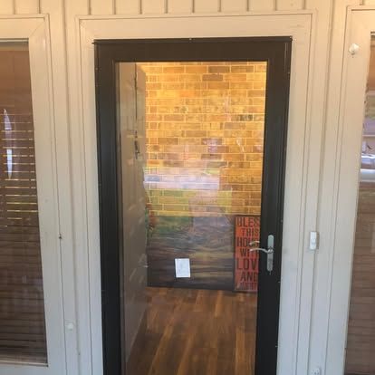 A black-framed glass storm door set into a light-colored wood panel wall, revealing a brick interior and wood floor.