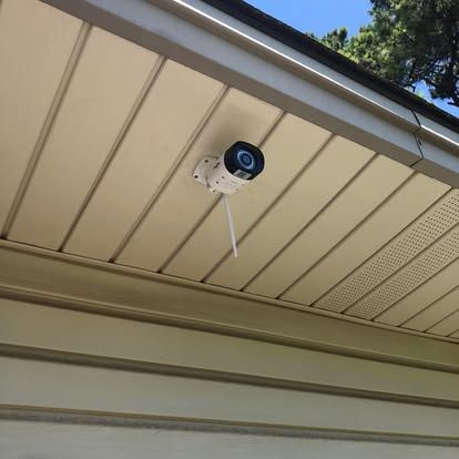 A white outdoor security camera with a protruding antenna is mounted to the underside of a beige house overhang.