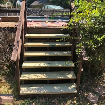 A set of new wooden stairs with dark brown railings leading up to a backyard pool deck on a sunny day.