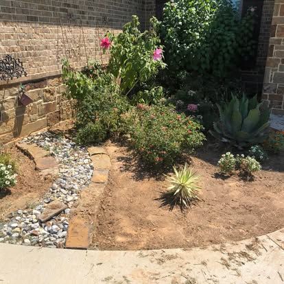 A flower bed with a stone-lined dry creek, small shrubs, blooming pink flowers, and a large succulent against a brick wall.