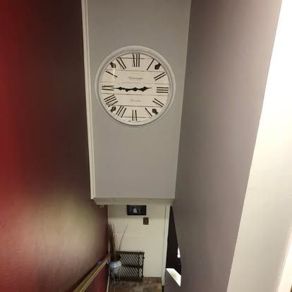 A large round wall clock with Roman numerals hangs on a light gray wall above a staircase landing with a metal rack.