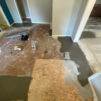 A home interior under renovation with patched concrete flooring, exposed subfloor, and construction materials on site.
