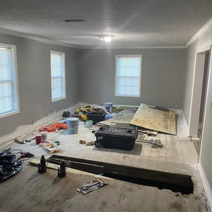 A room under renovation with gray walls, exposed subflooring, a hole in the floor, scattered tools, and building materials.