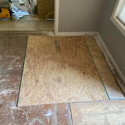 Newly installed plywood subflooring patches set into an unfinished room with gray walls and a visible tool in the background.