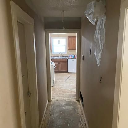 A narrow hallway with taupe walls leads toward a kitchen featuring brown cabinets and a white dishwasher.