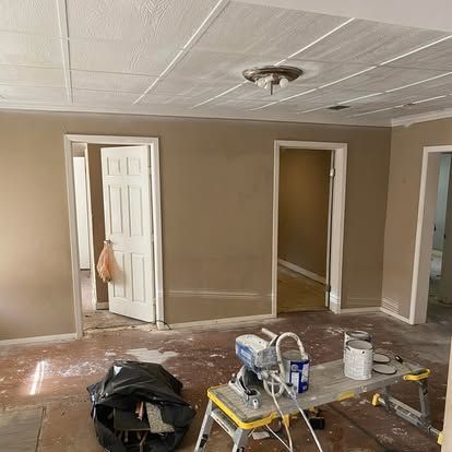 A room under renovation with beige walls, white trim, a paint sprayer, and a small folding table on a construction floor.