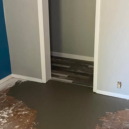 A room with gray walls and a doorway leading to wood-look flooring, with a section of floor covered in fresh gray concrete.