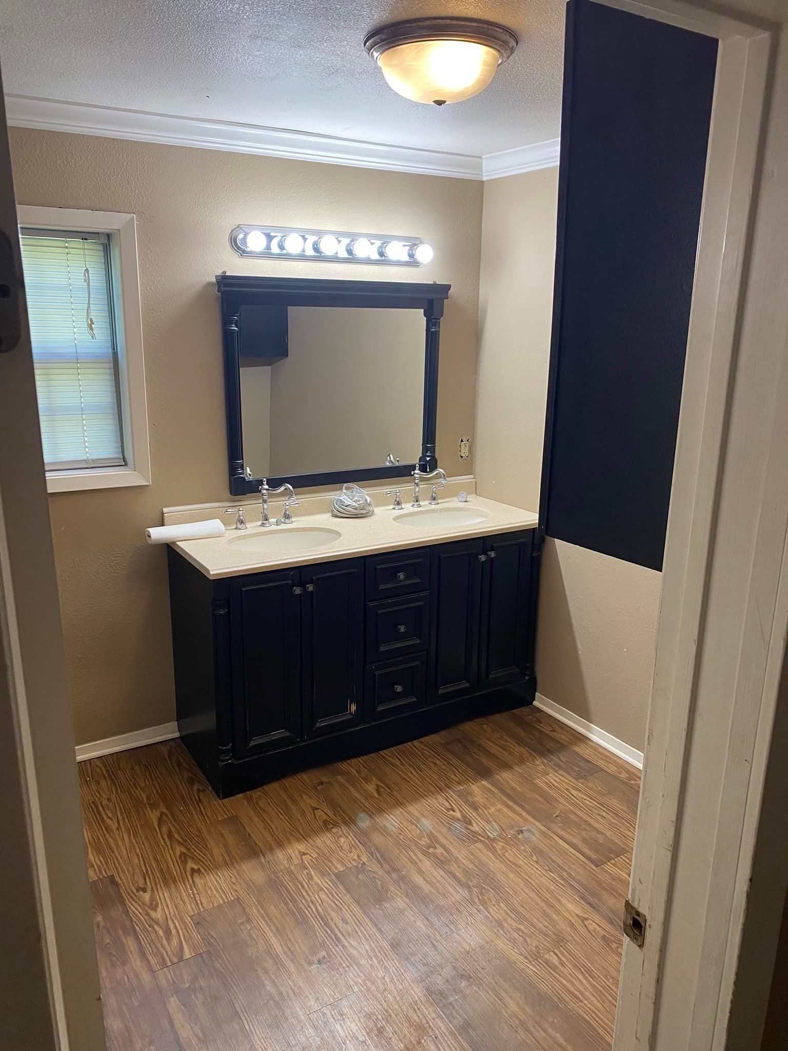 A double-sink bathroom vanity with dark cabinets, a framed mirror, and light countertop, set against tan walls.