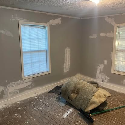 A room under renovation with gray walls, patched drywall spots, two windows with blinds, and a rolled-up rug on the floor.