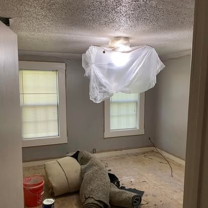 An unfinished room with gray walls, two windows, a light covered in plastic, and a rolled carpet on the floor.