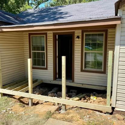 Construction of a wooden deck frame in front of a tan house with a door between two windows.