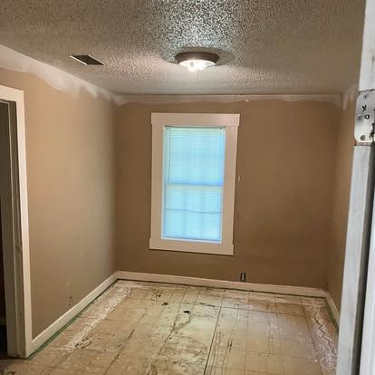 An empty room with tan walls, white trim, a textured ceiling, a center light fixture, and a subfloor ready for flooring.