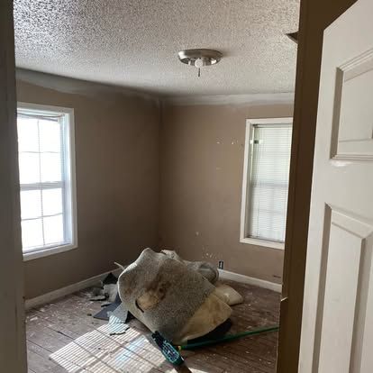 An empty room under renovation with tan walls, exposed wooden floors, two windows, and a rolled-up rug in the center.