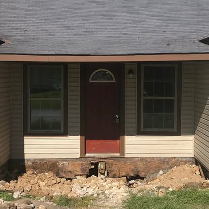 A house entrance with a red door and two windows above a foundation that is missing its porch steps and facing dirt.