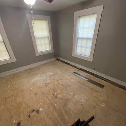 An empty bedroom with gray walls, two windows, and unfinished plywood subflooring.