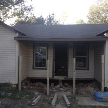 A house exterior with an unfinished porch, showing exposed foundation, dirt, and several vertical wooden support posts.