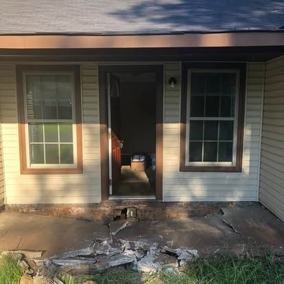A house entrance with light tan siding, two brown-framed windows, an open door, and a cracked, crumbling concrete porch.