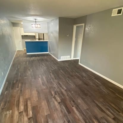 An empty room with dark wood-look flooring, light grey walls, a doorway, and a view into a kitchen with a blue island.