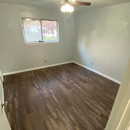 An empty bedroom with light gray walls, dark wood-look flooring, a window, and a ceiling fan light fixture.