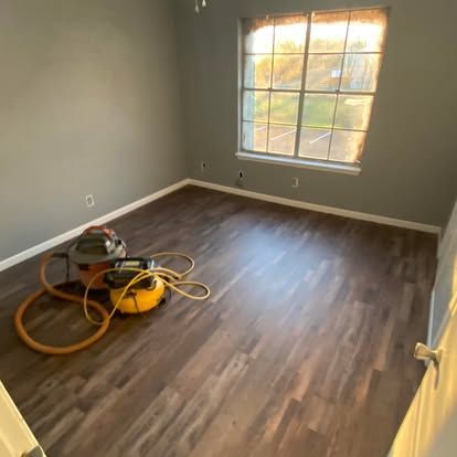A vacant bedroom with dark wood-look laminate flooring, grey walls, a single window, and a shop vacuum on the floor.