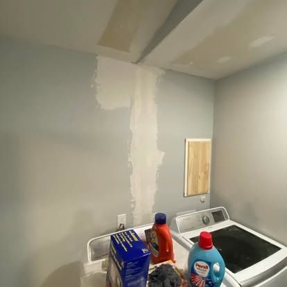 A laundry room wall with a vertical patch of white spackle and a washing machine with detergent bottles on top.