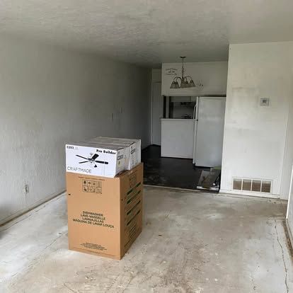 A large cardboard box sits on a bare concrete floor in a sparsely furnished, white-walled apartment living area.