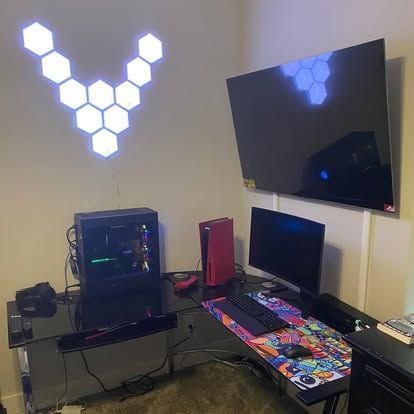 A desk setup with a gaming PC, red PlayStation 5, monitor, wall-mounted TV, and hexagonal light panels arranged in a V-shape.